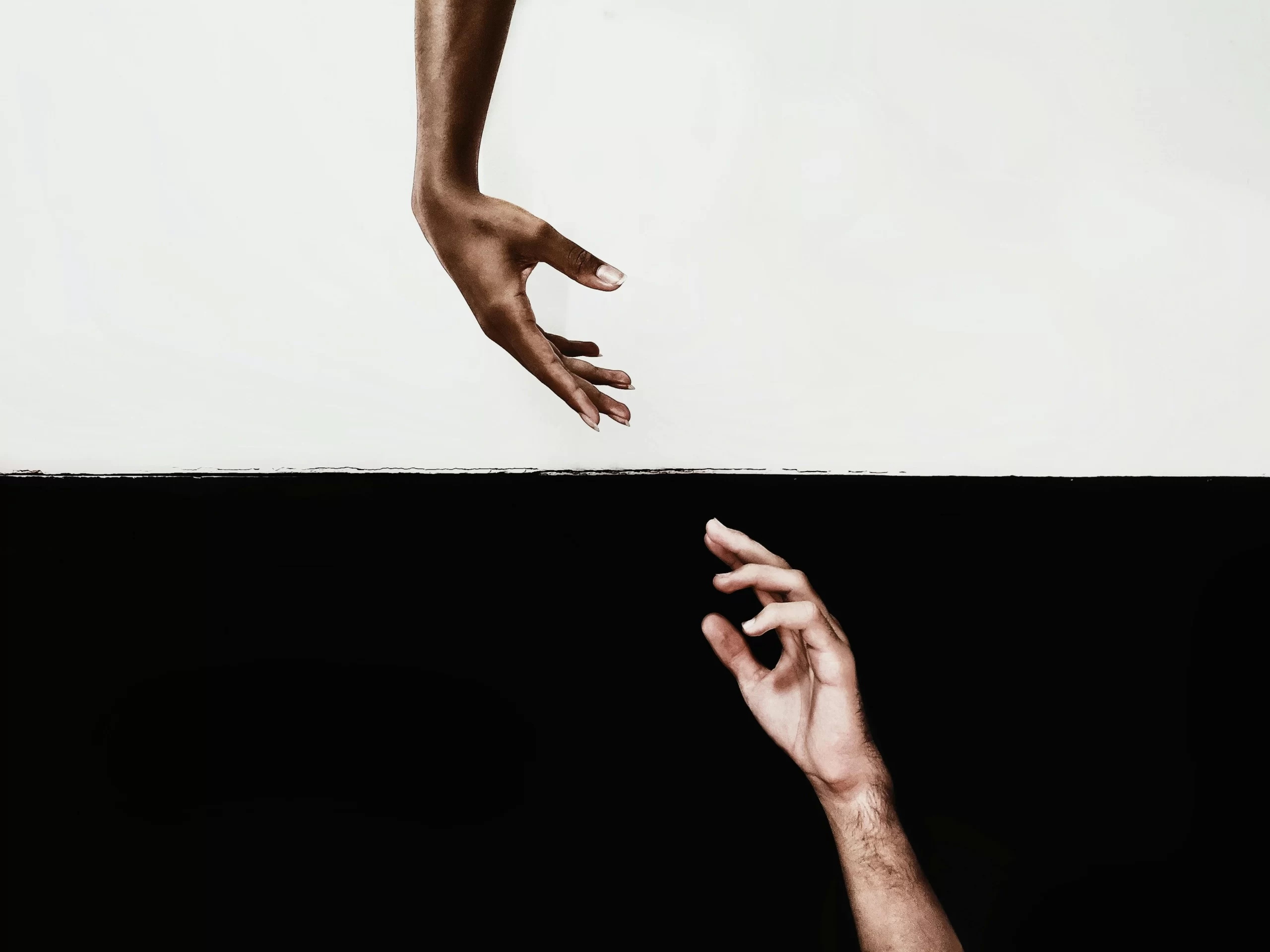 "Two hands reaching out to each other against a strikingly divided black and white background, symbolizing connection and contrast. The top hand is dark-skinned and gracefully extended downward, while the bottom hand is light-skinned, reaching upward, creating a visually compelling focal point that highlights unity and separation.